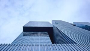 Low-angle view of several modern glass skyscrapers against a cloudy sky. The buildings feature sleek, reflective facades and sharp angles, embodying architectural strategy and complexity. The overall tone of the image is cool and contemporary.