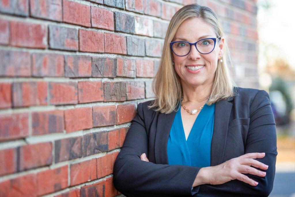 A woman with shoulder-length blonde hair and glasses stands confidently with her arms crossed, leaning against a brick wall. She is wearing a teal blouse and a black blazer, smiling at the camera. The background hints at an exterior setting, epitomizing leadership and strategy in her demeanor.