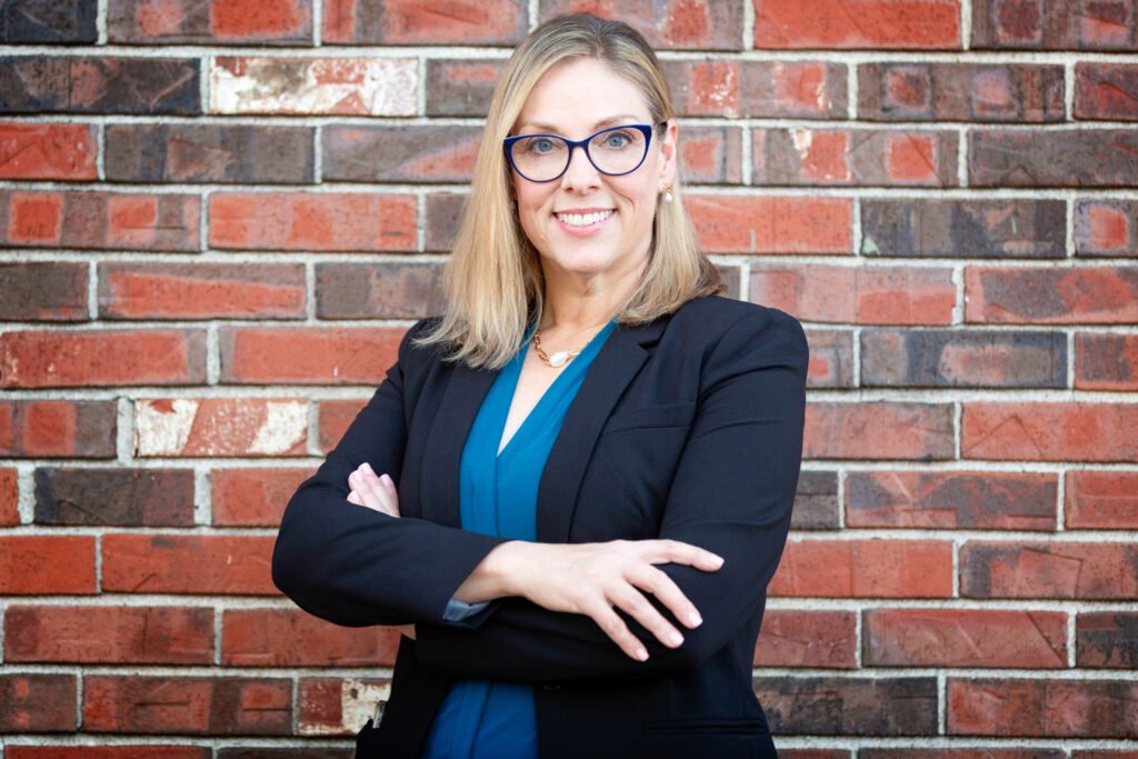 A confident woman with blonde hair and glasses stands with her arms crossed in front of a red brick wall. Wearing a blue top and black blazer, she smiles directly at the camera, exuding an air of coaching expertise and a focus on change management.