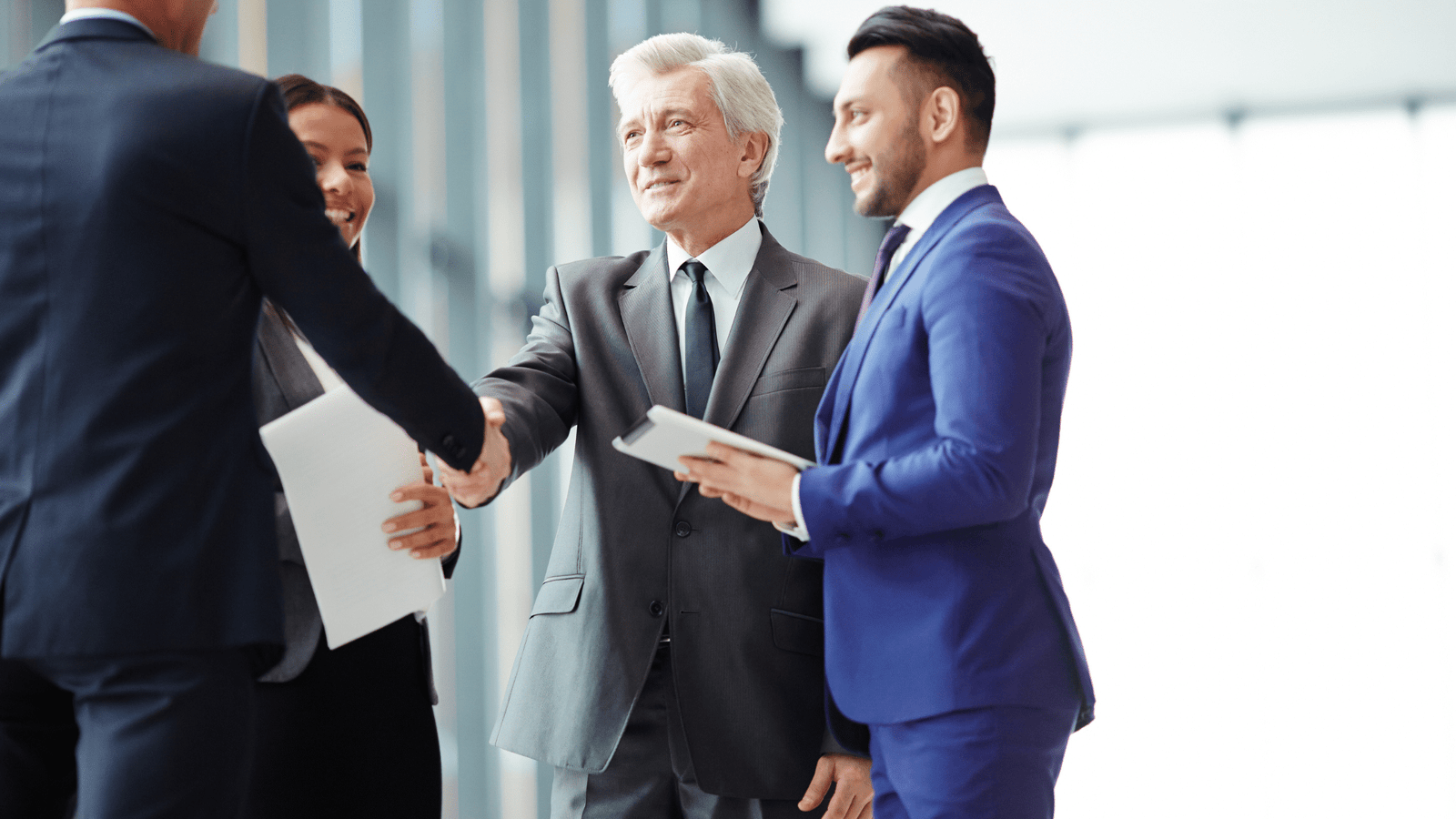 A group of business professionals, including a gray-haired man in a suit shaking hands with another person, stand together in a modern office setting. They are smiling and holding documents, indicating a successful meeting or agreement, showcasing impressive results from effective change management.
