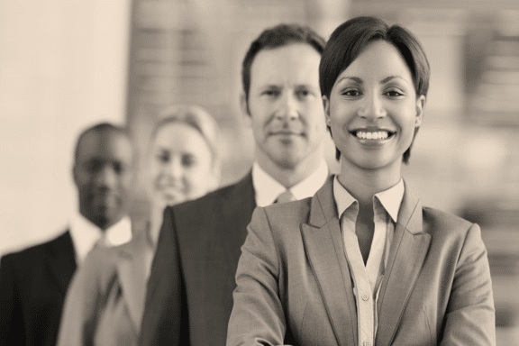 A black and white photo shows a group of four smiling business professionals in formal attire. The woman in the front is closest to the camera, embodying effective change management, with three people, two men and a woman, standing behind her in a blurred background, all looking forward.