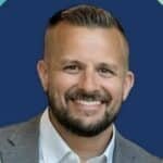 A smiling man with short, neatly styled hair and a trimmed beard, wearing a gray suit jacket and white shirt, stands in front of a solid blue background—perfect for consulting or technology professionals.