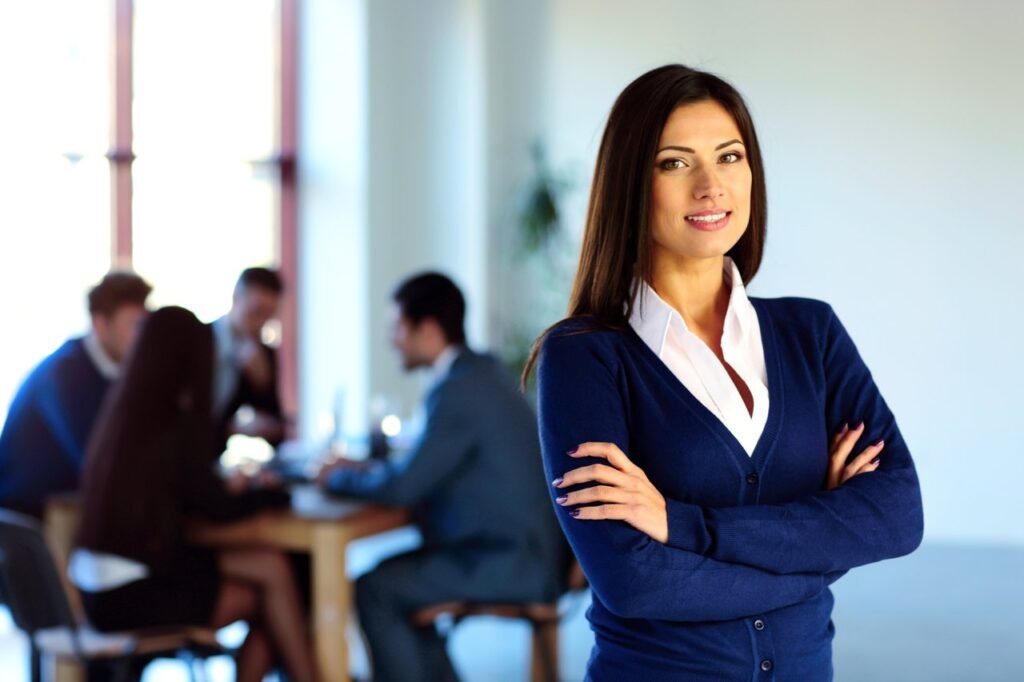 A confident woman in business attire stands with her arms crossed, exuding leadership while smiling at the camera. In the background, a group of four people sit around a table in a modern, well-lit office setting, engaged in strategy discussions. - Tapestry AG Woman Building Leadership Skills in Change Management