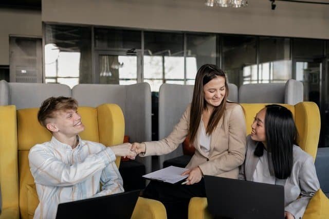 Three people are seated in a modern office space with laptops in front of them. A man in a white shirt is shaking hands with a smiling woman in a beige blazer holding documents, symbolizing strong leadership. Another woman in white smiles as she observes, reflecting effective change management. Yellow chairs and glass partitions are visible. - Tapestry AG One man and two women in a conference room discussing change management and strategic initative success.