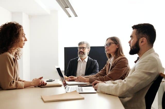 A group of four individuals is seated in a conference room. Three of them, comprising two men and one woman, sit on one side of the table with laptops in front of them, while a woman sits across from them. They appear to be engaged in a discussion about change management strategies.