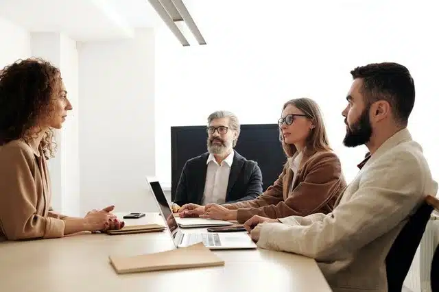 A group of four individuals is seated in a conference room. Three of them, comprising two men and one woman, sit on one side of the table with laptops in front of them, while a woman sits across from them. They appear to be engaged in a discussion about change management strategies.