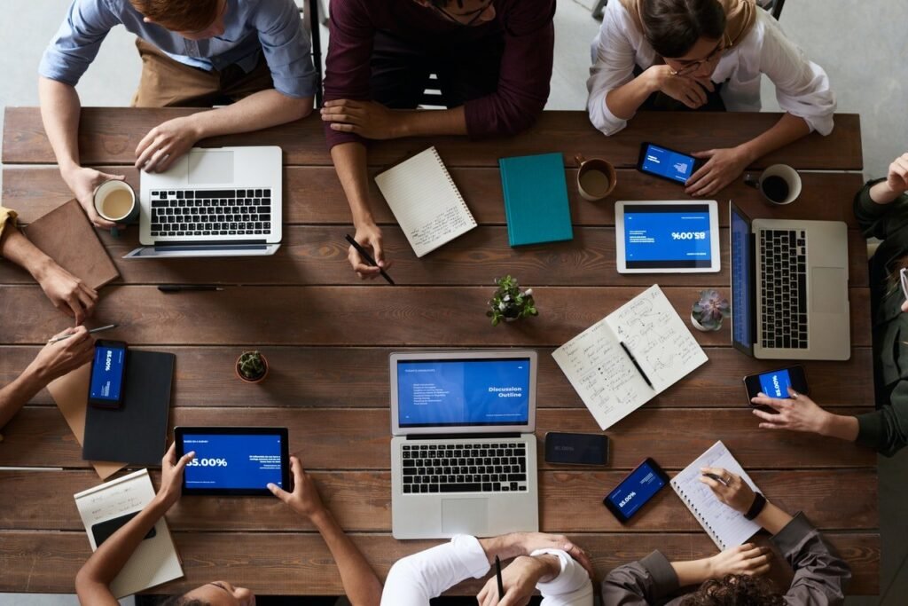 A group of people sitting at a wooden table, collaborating with laptops, tablets, notebooks, and smartphones. Papers, pens, coffee cups, and small potted plants are scattered on the table. The individuals seem to be engaged in a strategy session or meeting focused on achieving results.
