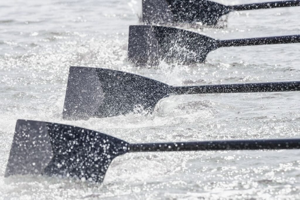 Close-up of four rowing oars in motion, splashing water as they move in unison. The paddles are black and the water appears to be choppy, indicating vigorous rowing. This scene captures the essence of teamwork and leadership, showcasing the results of effective coaching and synchronized effort.