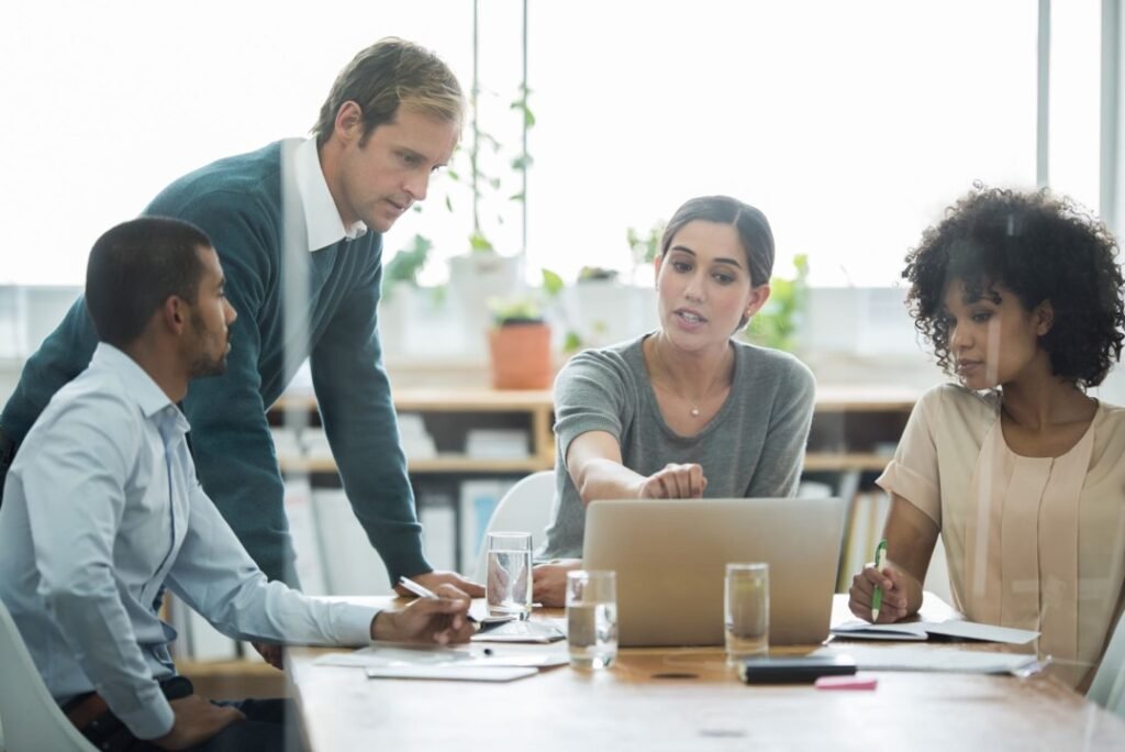A group of four people are seated around a wooden table, engaged in a discussion on strategy. One woman points at a laptop screen while the others look on attentively. The workspace is bright, with plants in the background and various office supplies on the table.