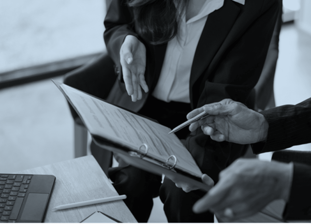 Two people in business attire are seated at a table, reviewing a document in a binder. One person is pointing at the papers with a pen, demonstrating effective leadership. A laptop is placed on the table beside them. The image, captured in black and white, underscores their focus on results.