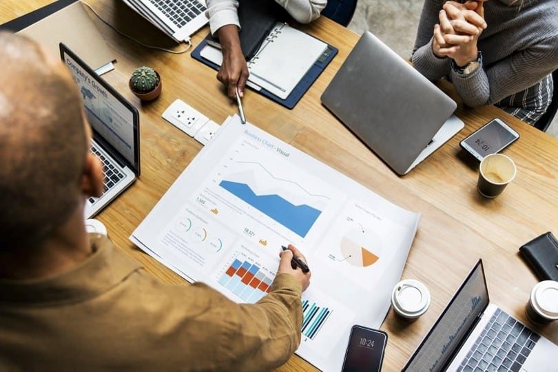A team is gathered around a table with open laptops, smartphones, coffee cups, a plant, and documents displaying charts and graphs. One person is pointing at a large sheet of paper with a pen, suggesting a strategy discussion or coaching session focused on driving results. - Tapestry AG A team is gathered around a table with open laptops, smartphones, coffee cups, a plant, and documents displaying charts and graphs. One person is pointing at a large sheet of paper with a pen, suggesting a strategy discussion or coaching session focused on driving results.