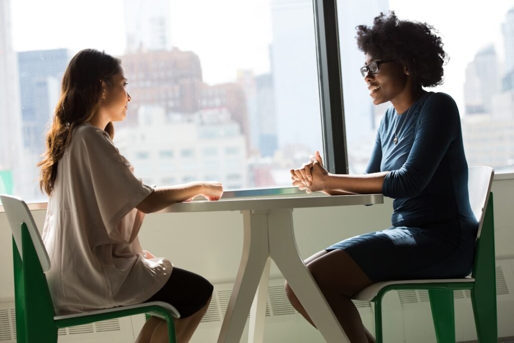 Two women sit across from each other at a table by a large window, engaged in conversation with a cityscape visible in the background.
