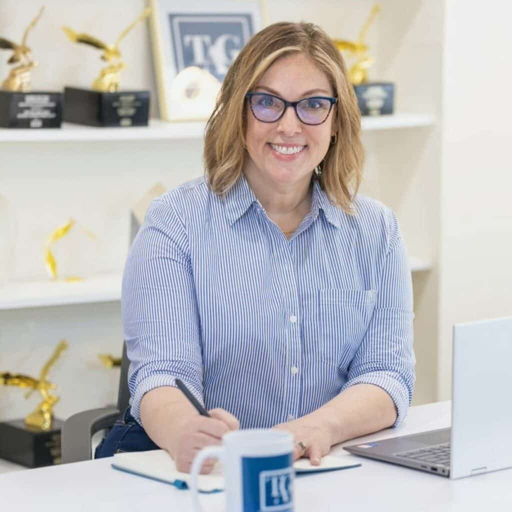 A woman with glasses and a striped shirt smiles while sitting at a desk, writing in a notebook. A laptop and branded mug are on the desk, reflecting her focus on coaching and alignment, with awards displayed on shelves in the background.