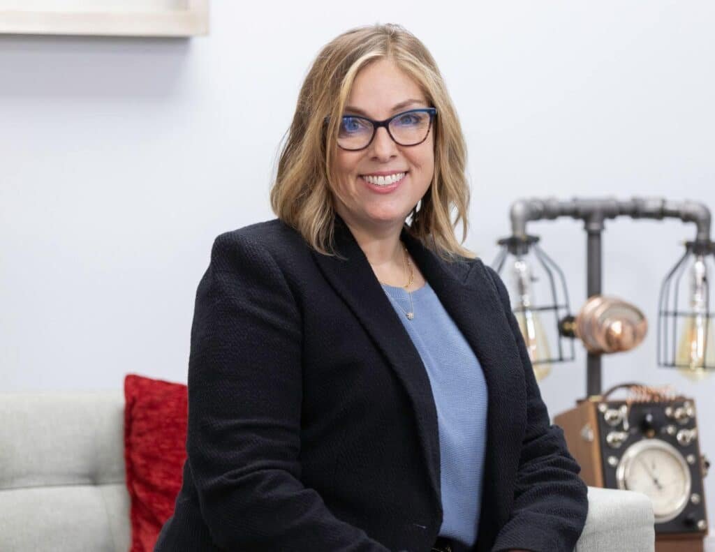A woman with shoulder-length blonde hair and glasses, wearing a navy blazer and blue top, sits on a light-colored couch. Behind her are a vintage telephone and industrial-style lamps. She is smiling at the camera. Mya Tyler A woman with shoulder-length blonde hair and glasses, wearing a navy blazer and blue top, sits on a light-colored couch. Behind her are a vintage telephone and industrial-style lamps. She is smiling at the camera.