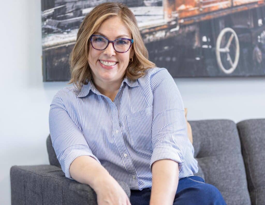 A woman with shoulder-length blond hair and glasses sits smiling on a gray couch. She is wearing a light blue striped shirt and navy pants. A painting or photograph featuring train wheels is visible in the background. - Tapestry AG A woman with shoulder-length blond hair and glasses sits smiling on a gray couch. She is wearing a light blue striped shirt and navy pants. A painting or photograph featuring train wheels is visible in the background.