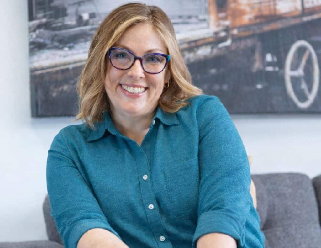 A smiling woman with shoulder-length blonde hair wearing glasses and a blue button-up shirt sits on a gray couch. A blurred black-and-white photo hangs on the wall behind her. A smiling woman with shoulder-length blonde hair wearing glasses and a blue button-up shirt sits on a gray couch. A blurred black-and-white photo hangs on the wall behind her.