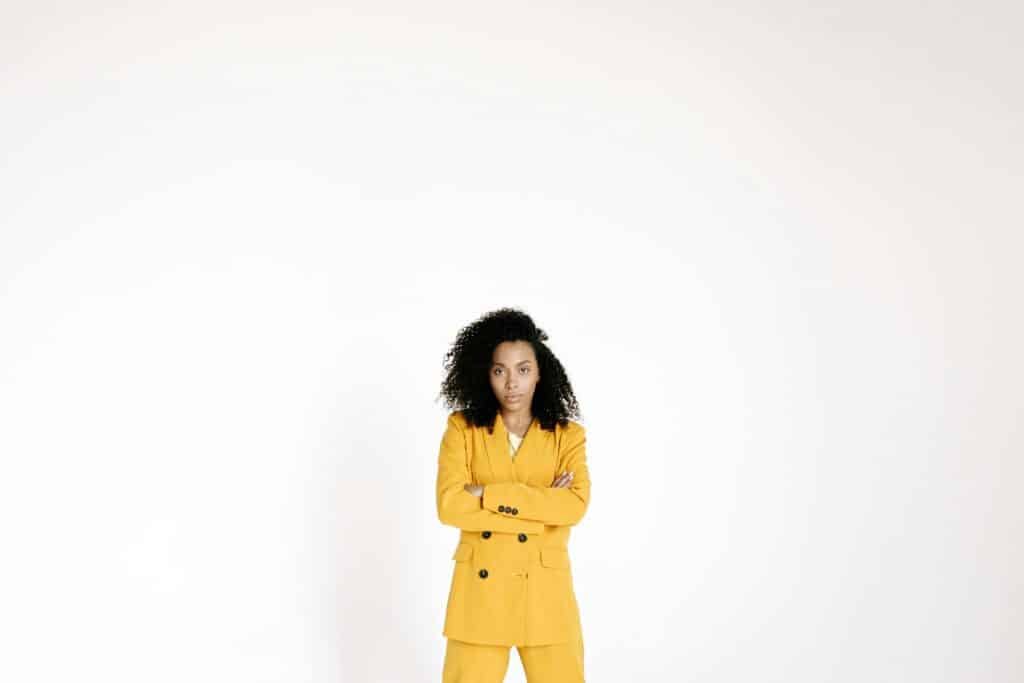 Portrait of a confident woman in a yellow suit with curly hair, arms crossed, posing in a studio setup.