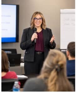 Female presenter in a dark blazer speaks into a handheld microphone in a conference room, with a screen and whiteboard in the background.