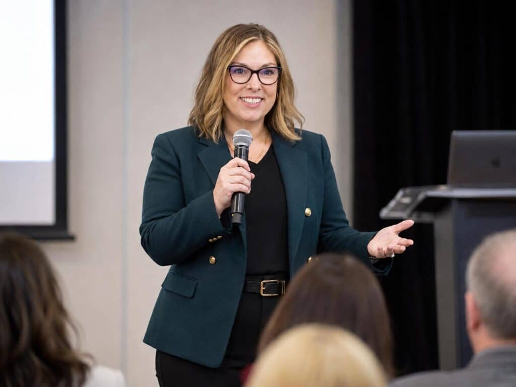 Woman with blond wavy shoulder length hair and glasses holding a microphone and speaking to a group.